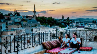 Yazın Goreme Kapadokya 'da, iki genç çatıda dinleniyor, içkilerini yudumlarken Türkiye' deki tarihi manzaranın nefes kesici manzarasını ve canlı günbatımını seyrediyor.