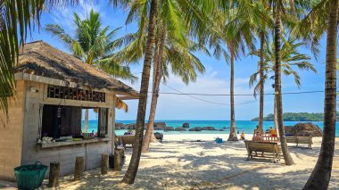 Sahil manzaralı sakin bir kulübe, uzun palmiye ağaçlarıyla çevrili, ziyaretçileri turkuaz okyanusun sakin bir gününün tadını çıkarmaya davet ediyor. Güneş banyosu ve yumuşak dalgalar huzurlu bir atmosfer yaratır..