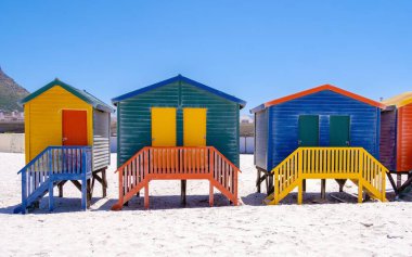 Cape Town 'daki muhteşem Muizenberg plajı boyunca uzanan, parlak boyalı plaj kulübeleri altın kum ve açık mavi gökyüzünün önünde duruyor..