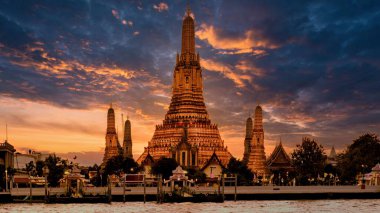 Alacakaranlık yaklaşırken, Wat Arun Bangkok 'ta çarpıcı bir gökyüzüne karşı dimdik ayakta duruyor, süslü dizayn edilmiş kuleler sıcak altın ve turuncu renkleri yansıtıyor. Ziyaretçiler nehir kenarında huzurlu bir anın tadını çıkarıyorlar..