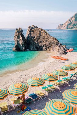 Ziyaretçiler, Monterosso al Mare 'deki güzel çakıl taşı plajında çizgili şemsiyeler altında renkli güneş pansiyonlarında dinleniyorlar. Berrak turkuaz su ve manzaralı uçurumlar İtalya 'da mükemmel bir yaz tatili kaçamağı yaratır.