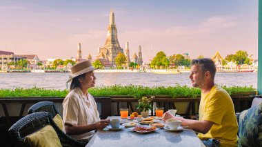 İki arkadaş Bangkok 'ta nehir kenarındaki terasta nefis bir kahvaltı yapıyorlar. Muhteşem Wat Arun Tapınağı' nın arka planında açık mavi bir gökyüzünün altında lezzetli yemeklerin tadını çıkarıyorlar..