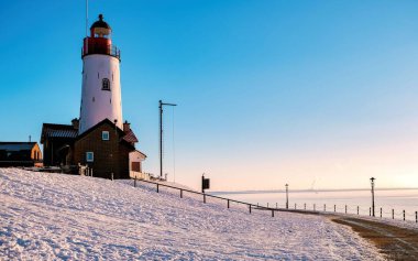 Kar, açık mavi bir gökyüzüne karşı dimdik duran pitoresk bir deniz fenerinin etrafındaki manzarayı kaplar. Sabah güneşi kıyı sahnesini aydınlatıyor, Urk, Hollanda