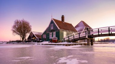 Resimli bir kış akşamında karla çevrili geleneksel bir Hollanda evi sergileniyor. Donmuş bir su yolu, günbatımının yumuşak renkleriyle davetkar bir yapıya götürür, Zaanse Schans, Hollanda
