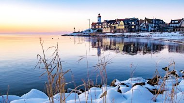 Sakin sular, karla kaplı kayaların çerçevelediği kış günbatımının pastel renklerini yansıtır. Alacakaranlıkta, Urk, Hollanda 'da güzel bir sahil manzarası yaratan antika evler ve deniz feneri hattı.