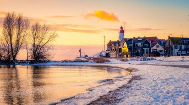 Resimli bir sahil köyü, büyüleyici bir deniz fenerinin yer aldığı kış günbatımının sıcacık parıltısıyla güneşleniyor. Hollanda 'da kıyı şeridi boyunca uzanan yapraksız ağaç siluetleri.