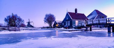 Kış Zaanse Schans 'a büyülü bir tılsım getiriyor, antika evler ve geleneksel yel değirmenleriyle. Donmuş ağaçlar Hollanda 'da alacakaranlıkta yumuşak bir pastel gökyüzünün altındaki donmuş kanalı çerçeveliyor.