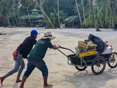 Koh Kood, Tayland, 13 Mart 2025, İki yerel işçi çöp dolu bir arabayı iterek kumsalda atık olarak kullandılar. 