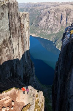 İki yürüyüşçü dramatik uçurumlarla çevrili bir fiyordun derin mavi sularına bakarken bir anlık korkunun tadını çıkarırlar. Kjeragbolten Lysefjord, Norveç