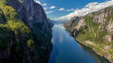 Norveç 'in Lysefjord kentindeki yüksek uçurumlar arasında çarpıcı bir fiyort uzanırken, yemyeşil alanlar kayalık kıyıları vurguluyor. Açık mavi su, canlı gökyüzünü yansıtır.