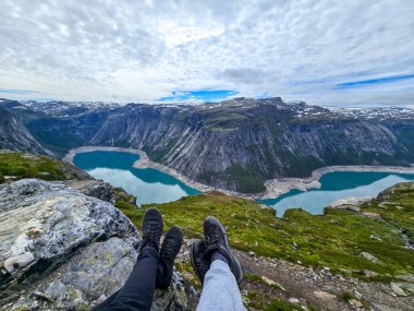 İki yürüyüşçü Norveç 'te nefes kesici bir fiyort manzarasına bakan kayalık bir zirvede dinleniyor. Durgun turkuaz sular, Norveç 'in dramatik dağ manzarasıyla tezat oluşturuyor.