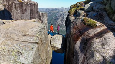 Canlı kıyafetler giymiş iki yürüyüşçü Norveç 'te çarpıcı fiyortlarla çevrili dramatik bir kaya oluşumunu keşfediyorlar. Aşağıda sakin mavi bir göl var, Kjeragbolten Lysefjord, Norveç