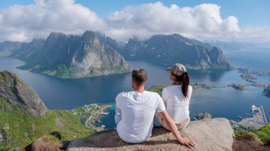 Bir çift, Norveç 'in Lofoten kentindeki muhteşem fiyort ve dağ manzarasına hayran kalarak bir kayanın üzerinde oturuyor. Açık mavi gökyüzü doğanın canlı renklerini güçlendirir. Macera aşıkları için mükemmel bir gün..