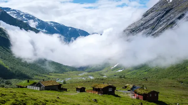 Görkemli dağlar, dinamik bir gökyüzünün altında geleneksel Norveç kulübeleriyle benekli sakin bir vadiyi kucaklıyor. Yeşillik ve yumuşak bulutlar barışçıl bir kırsal atmosfer yaratıyor..