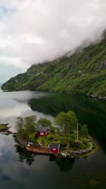 Norveç 'te, bulutlu bir gökyüzünün altındaki görkemli dağlarla çevrili sakin sularda yansıyan büyüleyici kırmızı kamaraları olan yemyeşil bir ada keşfedin. Lovrafjorden, Norveç