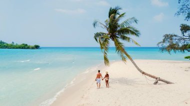 Huzurlu bir tropikal cennette, güneşli bir kumsal boyunca el ele yürüyen iki insan. Titreyen okyanusa doğru eğilen bir palmiye ağacı gibi hafif dalgalar ayaklarının dibinde döner..