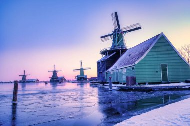 Rüzgâr değirmenleri güneş batarken buzlu suları yansıtıyor. Resimli bir Hollanda köyü olan Zaanse Schans 'ta. Kış renkleri, Hollanda 'nın zengin mirasıyla dolu büyüleyici bir sahneyi aydınlatıyor.