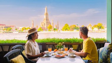 Çift Bangkok 'ta nehir kenarında huzurlu bir kahvaltı yapıyor, Wat Arun ve yemyeşil manzaralı canlı yemekler yiyor, açık bir gökyüzü altında mükemmel bir sabah kaçışı yaratıyor..