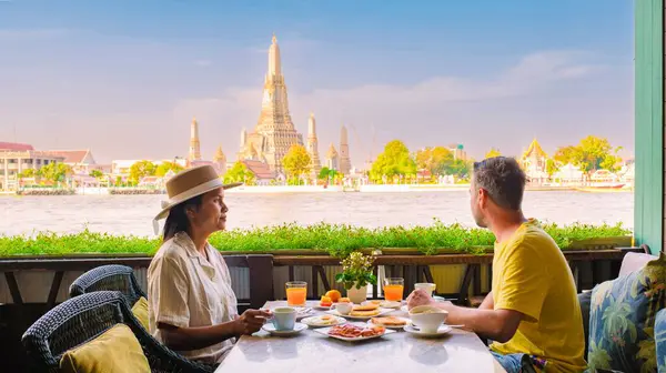 Çift Bangkok 'ta nehir kenarında huzurlu bir kahvaltı yapıyor, Wat Arun ve yemyeşil manzaralı canlı yemekler yiyor, açık bir gökyüzü altında mükemmel bir sabah kaçışı yaratıyor..