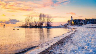 Batan güneşin altın rengi gölün sakin sularından yansıyarak karlı sahilde sıcak bir parıltı oluşturur. Urk Hollanda deniz feneri
