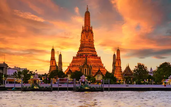 Alacakaranlık Bangkok 'u sararken Wat Arun altın ışıklarıyla görkemli bir şekilde aydınlanıyor. Ziyaretçiler, Chao Phraya Nehri 'nin sakin sularından yansıyan çarpıcı mimari ayrıntıların keyfini çıkarıyorlar..
