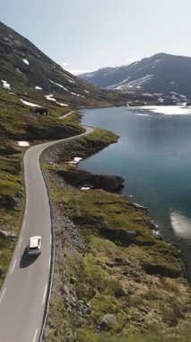 Norveç 'in nefes kesici güzelliğini keşfedin. Etrafı görkemli dağlarla çevrili sakin bir gölün yanındaki dolambaçlı yolda bir araba geziniyor. Dokunulmamış cazibe ve sükunet deneyimi..
