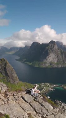 Kayalık kayalıkların tepesine tünemiş bir gezgin, Norveç 'in muhteşem dramatik fiyortlarının ve zümrüt vadilerinin tadını çıkarıyor. Canlı mavi su, yükselen dağları ve yuvarlanan bulutları yansıtır. Lofoten