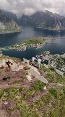 Norveç 'teki nefes kesen Lofoten Adaları' na bakan bir dağın kenarında iki yürüyüşçü duruyor. Huzurlu sular ve aşağıdaki köy görkemli dağların çerçevelediği görkemli bir manzara yaratır..