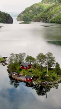 Norveç 'te sakin bir fiyort ile çevrelenmiş parlak kırmızı bir kulübe yemyeşil yemyeşil arasında huzur içinde duruyor. Sakin sular çarpıcı manzarayı yansıtıyor, Lovrafjorden, Norveç