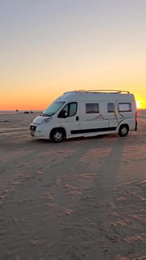 Akşam Romo Lakolk plajında güneş ufukta batarken yumuşak kumlara gömülmüş beyaz bir karavan sergileniyor. Danimarka 'nın bu güzel kıyı ortamında rahatlama ve macera birleşti