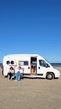 İki arkadaş, Romo Lakolk plajının kumlu kıyılarındaki bir karavanın dışındaki sandalyelerde dinleniyor. Açık mavi gökyüzü Danimarka 'daki bu sakin sahil çekilmesinin atmosferini yükseltiyor