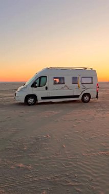 Danimarka 'nın Romo Lakolk plajının kumlu sahillerinde, çarpıcı bir günbatımına karşı bir karavan sergilenen huzurlu bir akşam yaşanıyor. Huzur, doğal güzelliklerle harmanlanır dalgalar yavaşça kıyıya vururken.