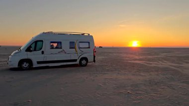 Güneş batarken Danimarka 'nın Romo Lakolk plajının geniş kumlarına park etmiş sakin bir karavan akşam gökyüzünde canlı renkler sergiliyor. Doğa güzelliği keşfi davet eder.