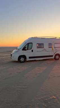 Gün batımında, Romo Lakolk Sahili 'nin kumlu sahiline park etmiş rahat bir karavan. Dalgalar, güneş doğanın üzerine sıcak bir parıltı saçarken nazikçe dolaşır ve sakin bir atmosfer yaratır..