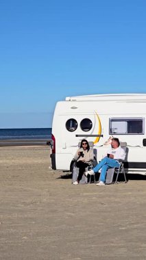 Danimarka 'daki Romo Lakolk plajındaki karavanın dışında birkaç kişi dinleniyor. Huzurlu kıyı manzarası, gevşemek ve doğal güzelliğin tadını çıkarmak için mükemmel bir zemin sunar..
