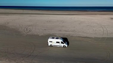 Danimarka 'daki Romo Lakolk plajının dingin güzelliğini deneyimleyin. Orada, açık mavi gökyüzünün altındaki kumlu geniş arazide yalnız bir karavan süzülüyor..