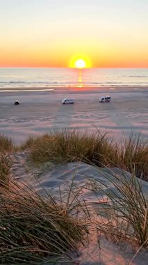 Gün batımında Danimarka 'daki Romo Lakolk Sahili' nin dingin güzelliğini görün. Gökyüzünün ılık renkleri sakin denizi yansıtırken araçlar geniş kumlu sahilde dinleniyor..