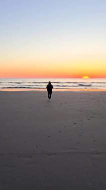 Danimarka 'nın Romo Lakolk plajında sakin bir akşam, sakin dalgalar ve canlı bir günbatımıyla çerçevelenmiş yumuşak kum üzerinde tek başına gezinen bir insan. Gökyüzü sıcak renklerle boyanmıştır..