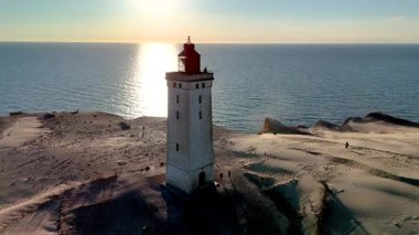 Günbatımı Danimarka 'nın Jutland kentindeki Rubjerg Knude Fyr Deniz Feneri' ne sıcak bir parıltı yayarak ikonik deniz feneri ve sakin kumulları vurguluyor. Ziyaretçiler kıyı şeridi boyunca yürüyüp barışçıl bir atmosfer yaratıyorlar.