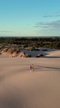 Danimarka 'nın Jutland kentindeki geniş kum tepeleri boyunca yürüyen iki kişi, canlı yeşillik ve dingin doğal güzelliklerle çevrili. Alacakaranlığın yumuşak ışığı nazikçe yollarını aydınlatır. Rabjerg Mil kum tepeleri