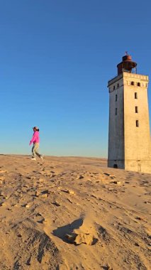 Günbatımında açık mavi gökyüzüne karşı duran tarihi deniz feneriyle Danimarka 'nın Jutland kentinin altın kum tepelerinde gezinen yalnız bir figür Rubjerg Knude deniz feneri