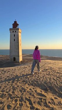 Genç bir gezgin, Jutland 'daki tarihi Rubjerg Knude Deniz Feneri yakınlarındaki kum tepeleri boyunca yürüyor. Günbatımının ılık renkleri manzarayı aydınlatır ve sakin bir atmosfer yaratır..