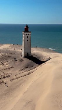 Rubjerg Knude Fyr Deniz Feneri kıyılarına doğru hafif dalgalar eserken ziyaretçiler sıcak güneşin tadını çıkarıyorlar. Danimarka 'nın Jutland şehrinde çarpıcı bir deniz feneri mavi gökyüzüne karşı dimdik duruyor.