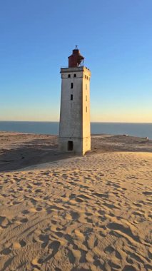 Danimarka 'nın Jutland kentinin sakin manzarasını keşfedin. Tarihi bir deniz feneri Rubjerg Knude altın kumların ve sakin bir günbatımının önünde dimdik durur.. 