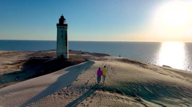 Danimarka 'nın Jutland kentindeki Rubjerg Knude Fyr Deniz Feneri' nin pitoresk kumları boyunca gezinen bir çift, sakin denize bakan tarihi bir deniz fenerinin yanında sıcak günbatımının keyfini çıkarırken,
