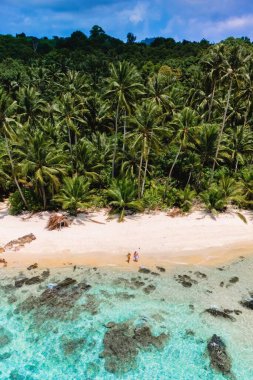 İki kişi canlı palmiyeler ve berrak turkuaz sularla çevrili el değmemiş kumlu bir plajda geziniyor. Koh Kood Tayland adasında tatilde olan çift.