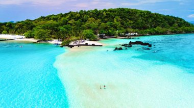 Turkuaz sular, iki bireyin sıcak güneşin tadını çıkardığı sakin bir kıyı şeridine nazikçe sarılır. Arkasından yemyeşil dağlar yükseliyor Koh Mak Tayland yakınlarındaki Koh Kham Adası.