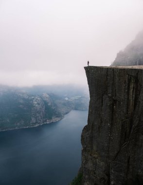 Kayalık bir uçurumun kenarında duran yalnız bir figür Preikestolen Norveç 'teki muhteşem fiyort görüntülerine bakıyor. Sisli manzara sakin bir atmosfer yaratır. Macera arayanlar için mükemmel. 