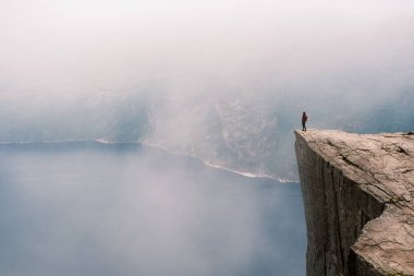 Preikestolen Norveç 'teki dramatik bir uçurumun kenarında duran yalnız bir maceracı sisli fiyorda ve aşağıdaki çarpıcı manzaraya tepeden bakıyor. Atmosfer sakin, merak uyandırıyor. 