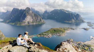 İki yürüyüşçü, Norveç 'teki Lofoten Adaları' nın göz kamaştırıcı manzarasına hayran kalarak kayalık bir çıkıntıda oturuyor. Canlı mavi sular, yükselen dağlar ve güzel köyler barışçıl bir atmosfer yaratıyor..